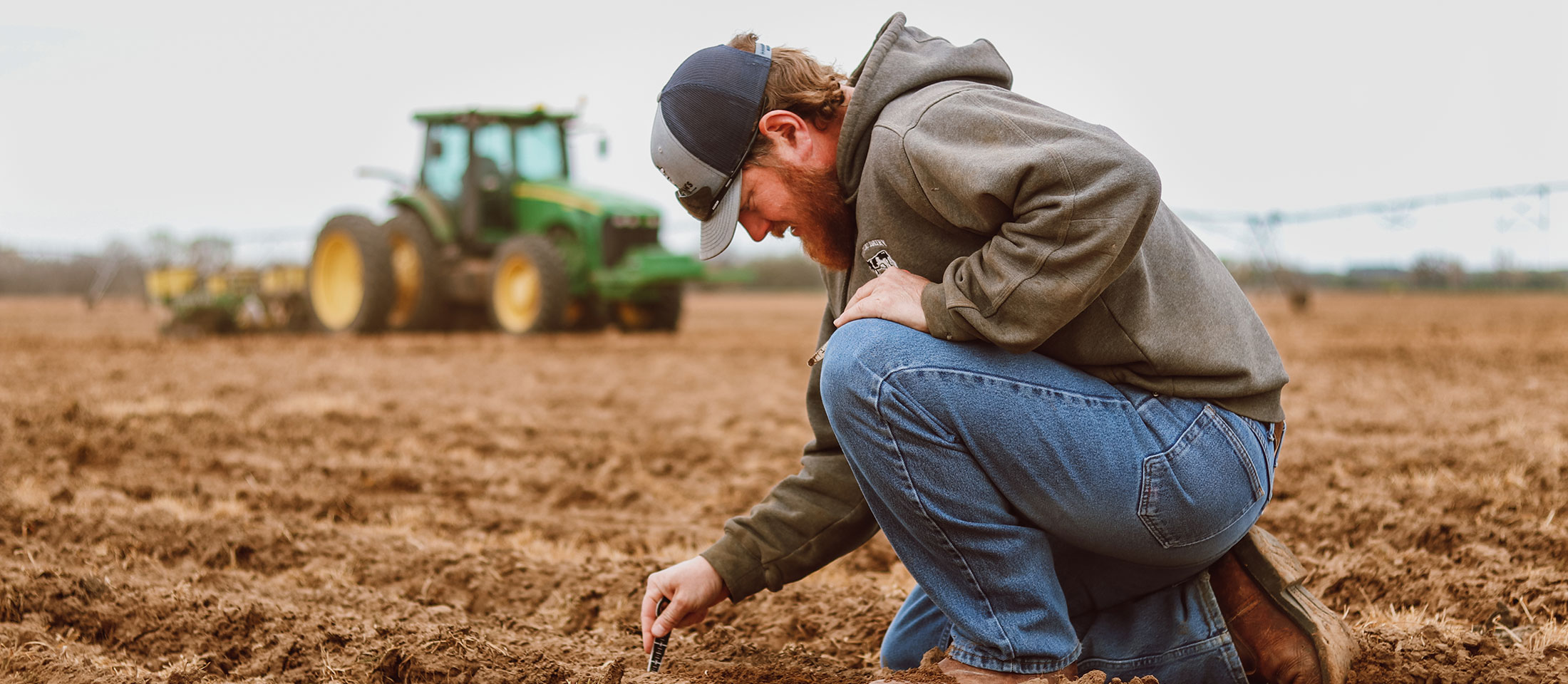 A man with facial hair wearing blue jeans, a gray hat, and a sweatshirt leaning down, looking at the ground, checking seed depth on their farm.